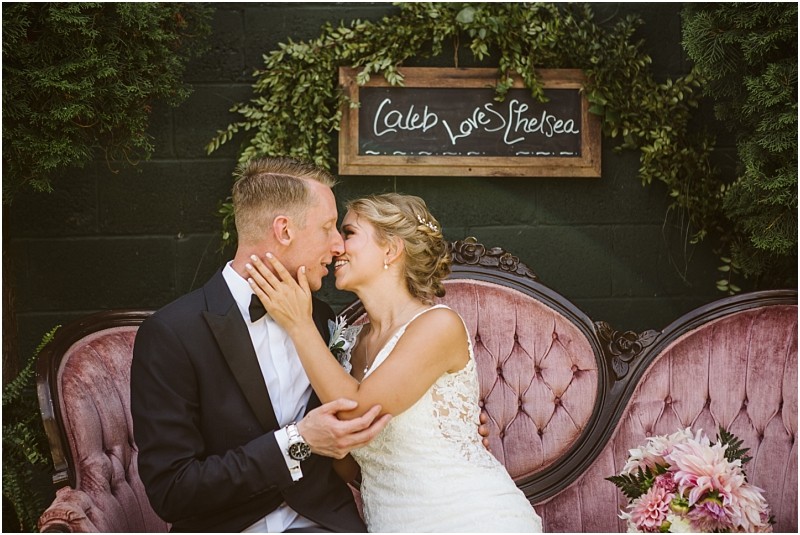 A bride and groom sit on a pink vintage sofa in snohomish smiling and embracing in front of a chalkboard sign that reads caleb loves chelsea surrounded by lush greenery and flowers Snohomish wedding photography