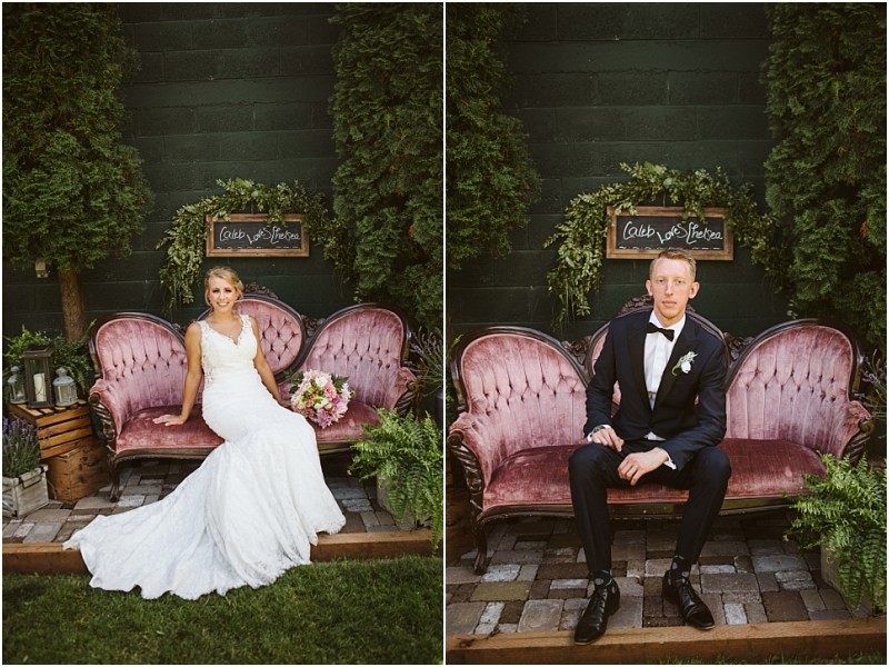 A bride in a white gown and a groom in a black tuxedo are each seated separately on a vintage pink sofa outdoors in snohomish with greenery and a chalkboard sign reading caleb & steven behind them Snohomish wedding photography