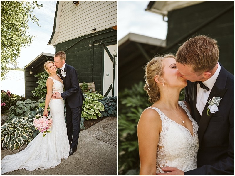 A bride in a white lace gown and a groom in a dark suit share a joyful embrace and kiss outdoors near leafy plants and a rustic snohomish building The bride holds a bouquet of pink flowers Snohomish wedding photography