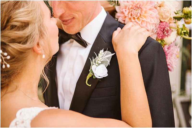 A bride and groom embrace closely in snohomish the brides arms around the grooms neck He wears a black tuxedo with a white boutonniere while her hair is styled with pearls Pink flowers brighten the background Snohomish wedding photography