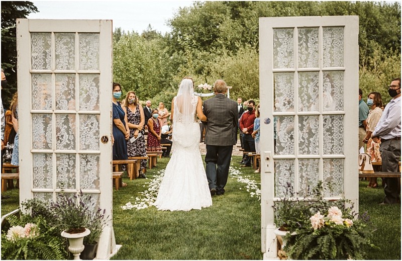 A bride in a white gown walks down an outdoor aisle with an older man flanked by guests on either side They enter through vintage glass doors decorated with lace surrounded by snohomish greenery and flowers Snohomish wedding photography