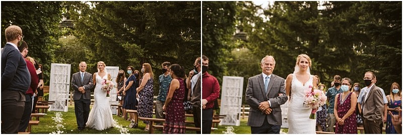 A bride in a white gown walks down an outdoor snohomish aisle with an older man surrounded by standing guests some wearing masks Trees and wedding decorations frame the scene Snohomish wedding photography