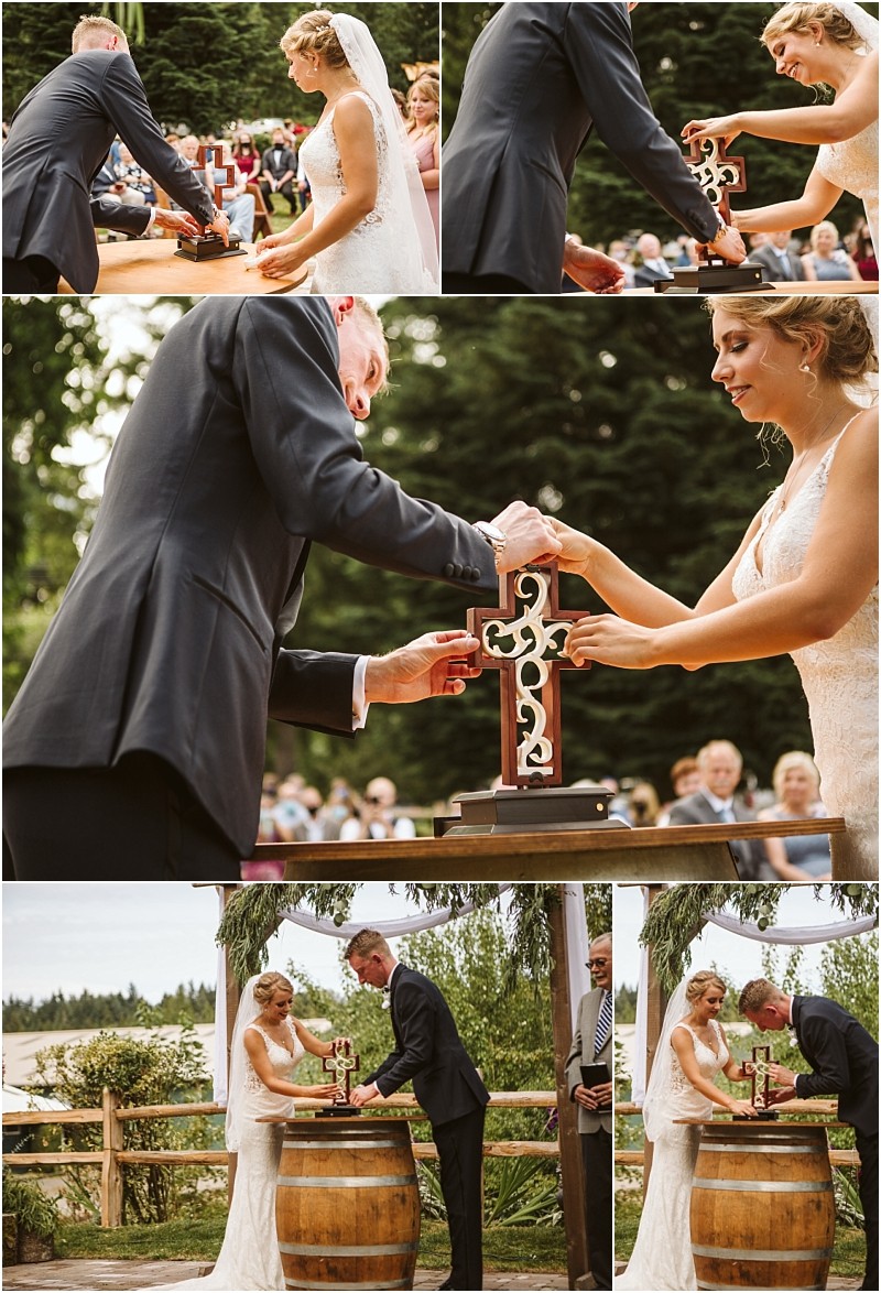 A bride and groom stand outdoors at a wooden barrel in snohomish assembling a decorative cross during their wedding ceremony surrounded by guests and trees Snohomish wedding photography