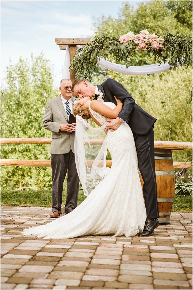 A groom in a dark suit dips his bride in a white lace gown for a kiss at their outdoor snohomish wedding ceremony while an officiant stands smiling behind them under a floral arch Snohomish wedding photography