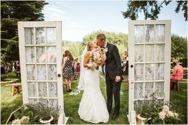 A bride and groom kiss outdoors between two vintage doors decorated with lace at a charming snohomish venue surrounded by wedding guests seated on benches and green trees in the background Snohomish wedding photography