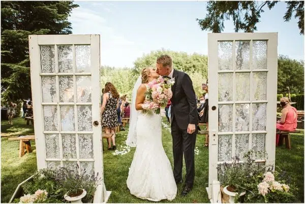A bride and groom kiss outdoors between two vintage doors decorated with lace at a charming Snohomish venue, surrounded by wedding guests seated on benches and green trees in the background. Snohomish Wedding Photography