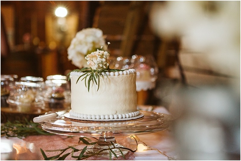 A simple white frosted cake decorated with small white flowers and greenery sits on a glass cake stand surrounded by jars and soft rustic wooden decor in a warmly lit snohomish room Snohomish wedding photography