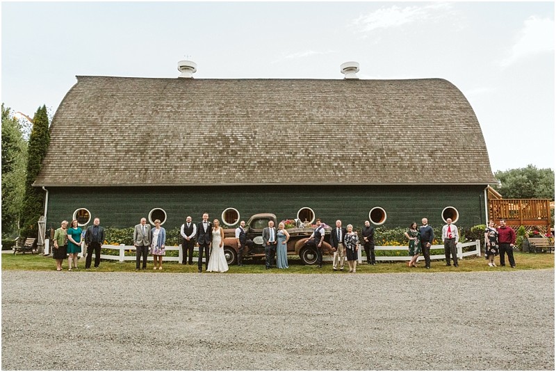 A wedding party stands in a line spaced apart in front of a large green barn with round windows and an old pickup truck in snohomish under a partly cloudy sky Snohomish wedding photography