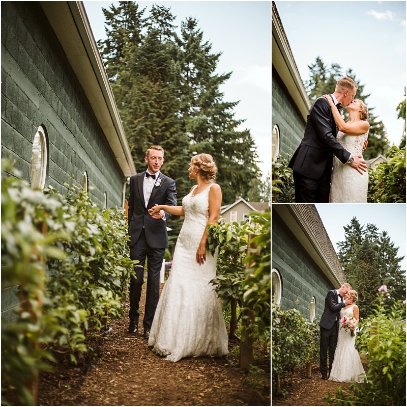 A bride in a white gown and a groom in a dark suit walk hold hands and share a kiss outdoors beside a green building surrounded by lush greenery in scenic snohomish Snohomish wedding photography