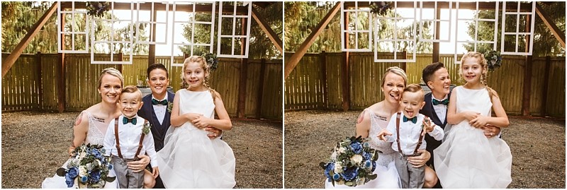A bride in a white dress and two children a boy in suspenders and a girl in a white dress pose and smile together in an outdoor wooden structure with hanging window frames in scenic snohomish Snohomish wedding photography