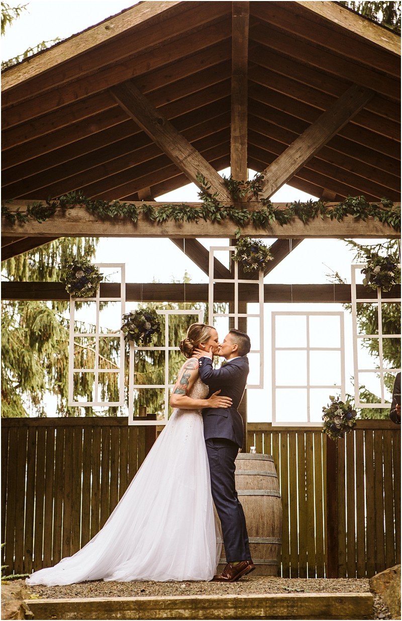 A bride and groom share a kiss under a wooden pavilion decorated with hanging window frames and greenery during their outdoor snohomish wedding ceremony Snohomish wedding photography