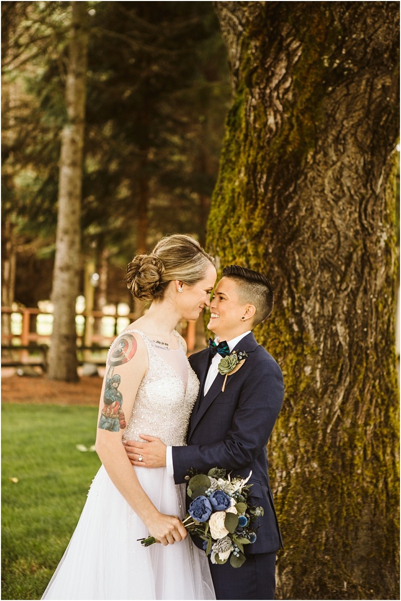 Two people in wedding attire stand close together smiling lovingly at each other under a large snohomish tree One holds a bouquet of blue and white flowers both appear happy and joyful on their wedding day Snohomish wedding photography
