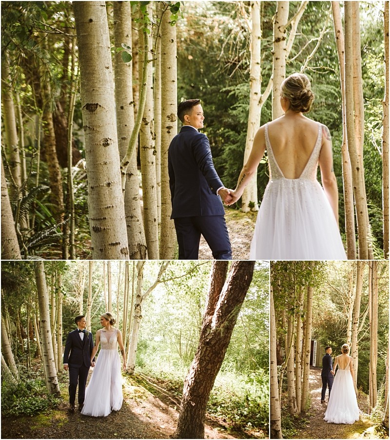 A bride and groom walk hand in hand through a sunlit snohomish forest The bride wears a white dress with an open back the groom is in a dark suit They stroll together along a wooded path surrounded by tall slender trees Snohomish wedding photography