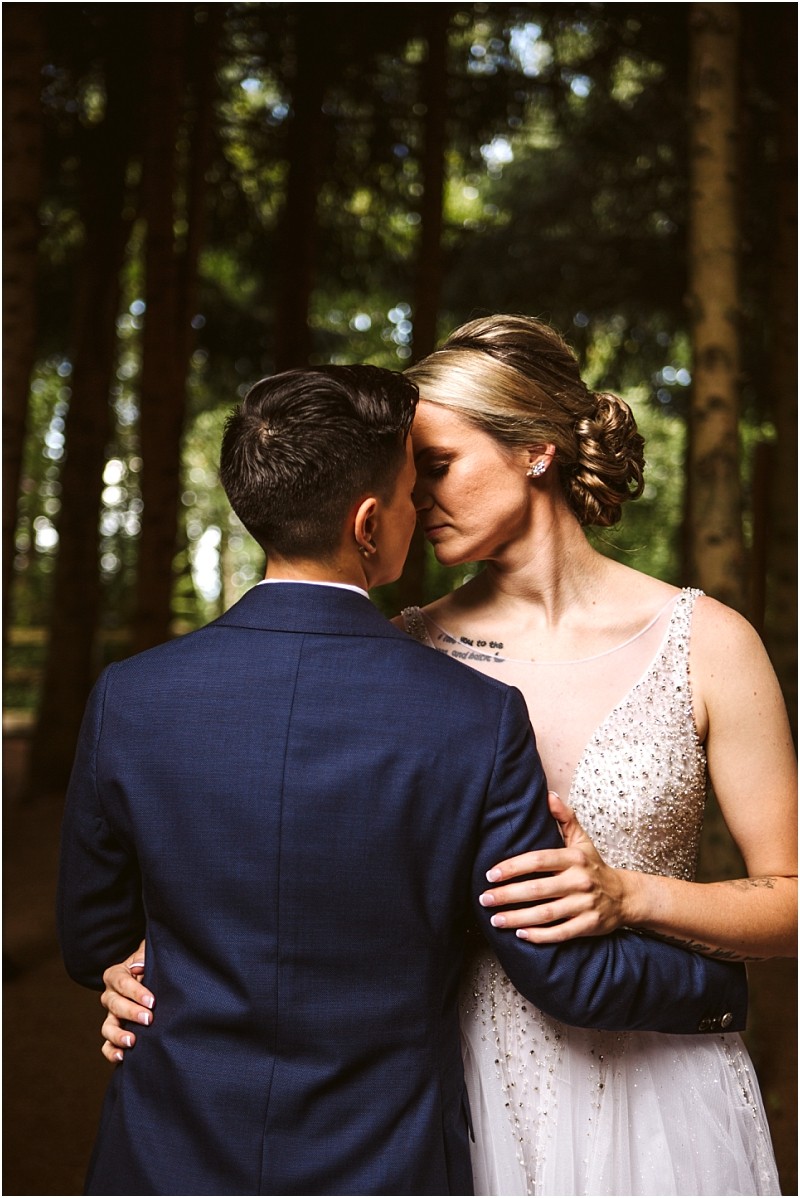 Two people in wedding attire embrace closely in a snohomish forest with their foreheads touching One wears a dark blue suit the other a white beaded dress Sunlight filters gently through the trees behind them Snohomish wedding photography