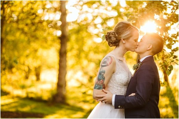 A couple dressed in wedding attire shares a kiss outdoors in snohomish with sunlight streaming through trees The person in the dress has visible arm tattoos adding personality to the warm golden scene Snohomish wedding photography
