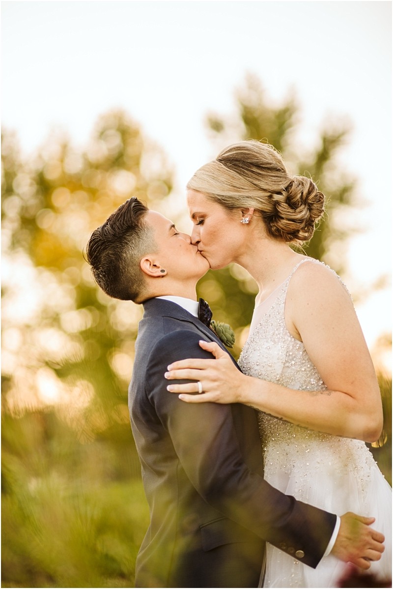 Two brides share a kiss outdoors on their snohomish wedding day one in a dark suit and the other in a white beaded gown with her hair up Sunlight and trees create a soft warm background Snohomish wedding photography