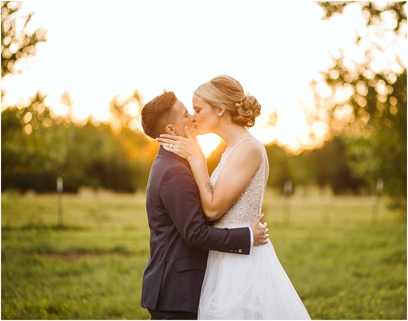 A couple in wedding attire shares a kiss outdoors at sunset surrounded by lush snohomish greenery The person on the left wears a dark suit while the person on the right wears a white wedding dress Snohomish wedding photography