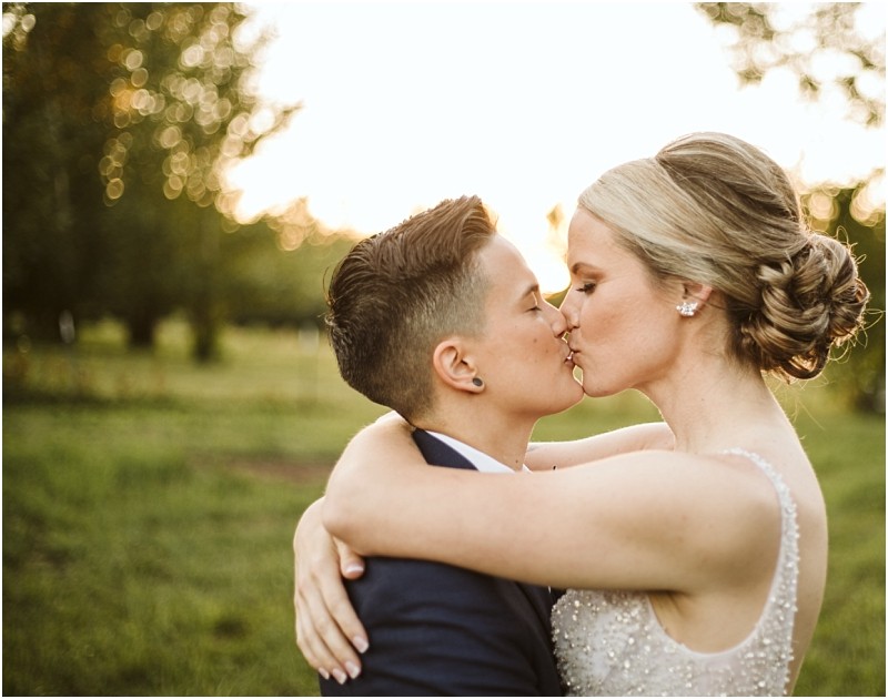 Two women embrace and kiss outdoors at sunset in snohomish one with short hair in a suit and the other with long hair in an updo and a beaded dress surrounded by trees and soft sunlight Snohomish wedding photography