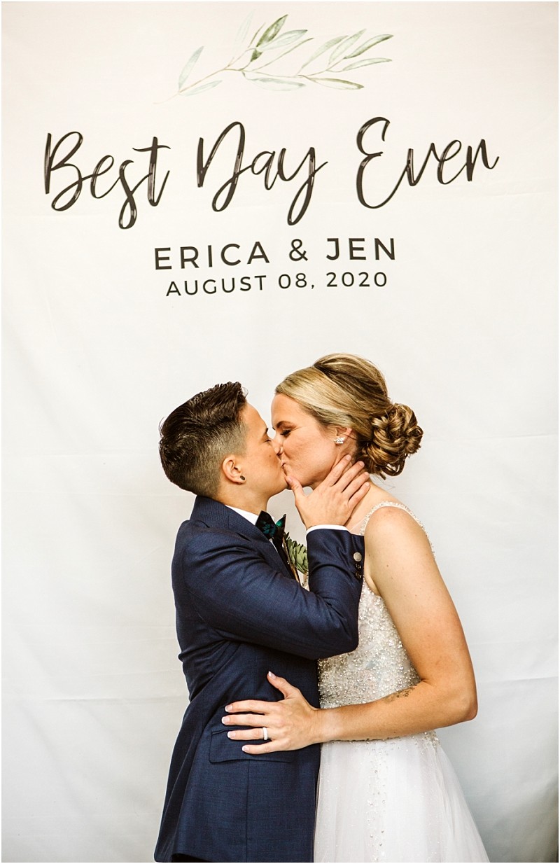 Two brides share a kiss in front of a white backdrop that reads best day ever erica & jen august 08 2020  one wears a suit and the other a wedding dress both look joyful and loving at their snohomish celebration Snohomish wedding photography