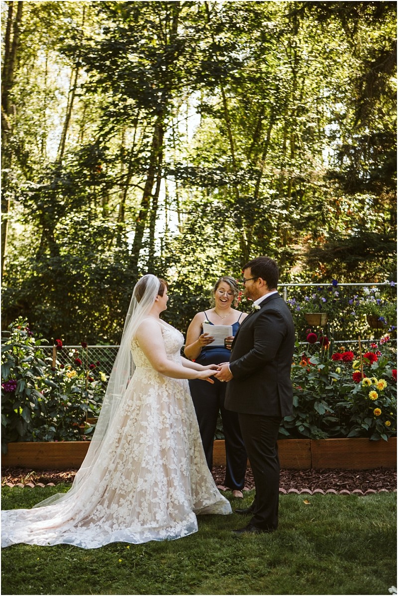A bride and groom stand holding hands outdoors during their snohomish wedding ceremony as an officiant reads from a book They are surrounded by green trees and bright flowers in a beautiful garden setting Snohomish wedding photography