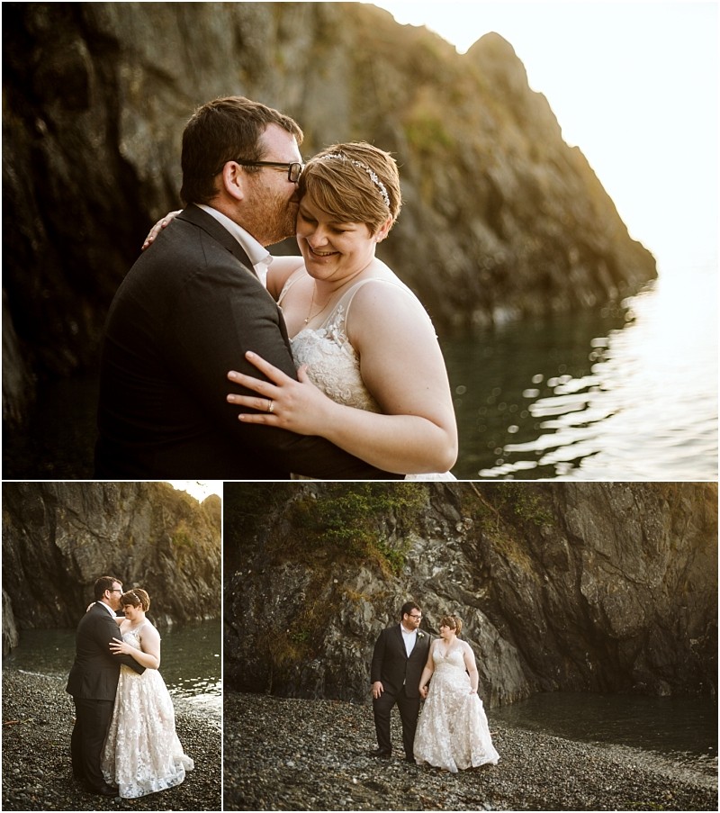 A couple in wedding attire embraces and smiles by a rocky snohomish shore at sunset The groom kisses the brides head they hold hands on the pebbled beach with cliffs and water in the background Snohomish wedding photography