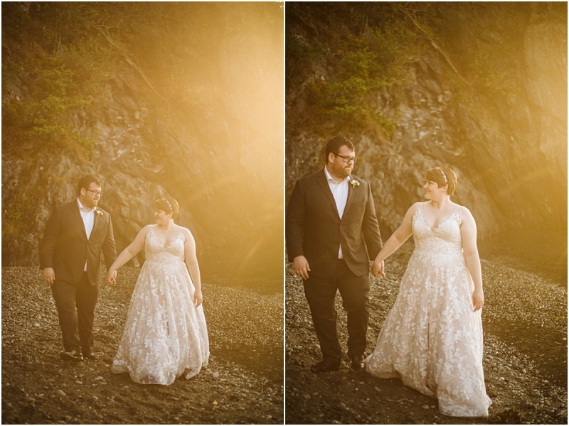 A couple dressed in a wedding gown and suit walk hand in hand on a rocky snohomish beach at sunset surrounded by warm golden light and a rocky hillside in the background Snohomish wedding photography