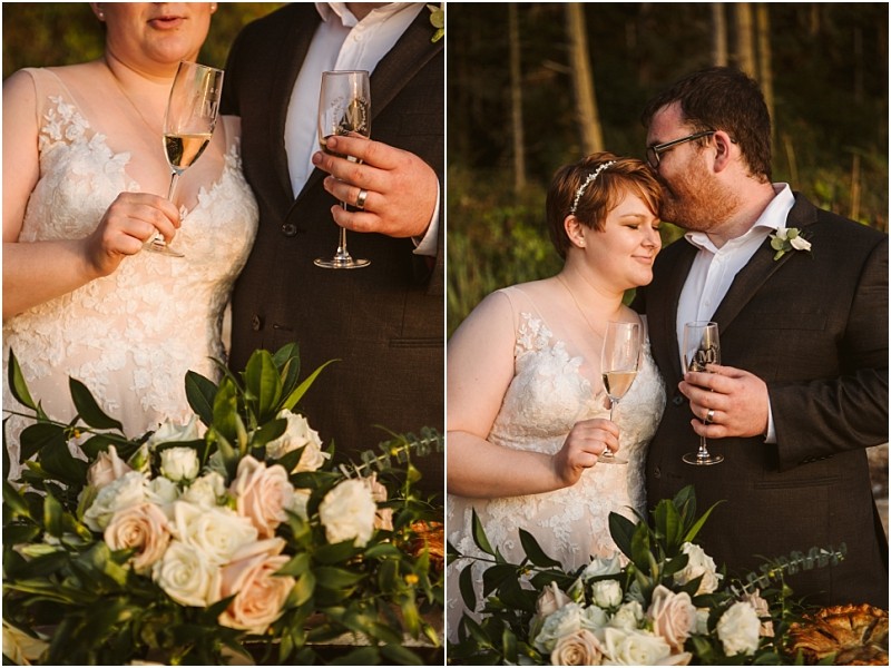 A bride and groom stand close together outdoors in snohomish holding champagne glasses The bride wears a lace dress and the groom a dark suit In front of them is a large bouquet of white and blush flowers with greenery Snohomish wedding photography