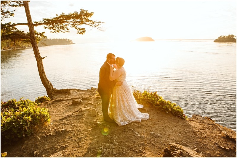 A couple embraces on a rocky cliff overlooking a calm body of water at sunset in snohomish with golden sunlight illuminating the scene and a tree leaning over the edge Snohomish wedding photography
