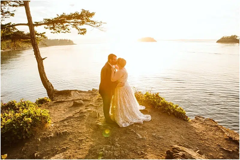 A couple embraces on a rocky cliff overlooking a calm body of water at sunset in Snohomish, with golden sunlight illuminating the scene and a tree leaning over the edge. Snohomish Wedding Photography