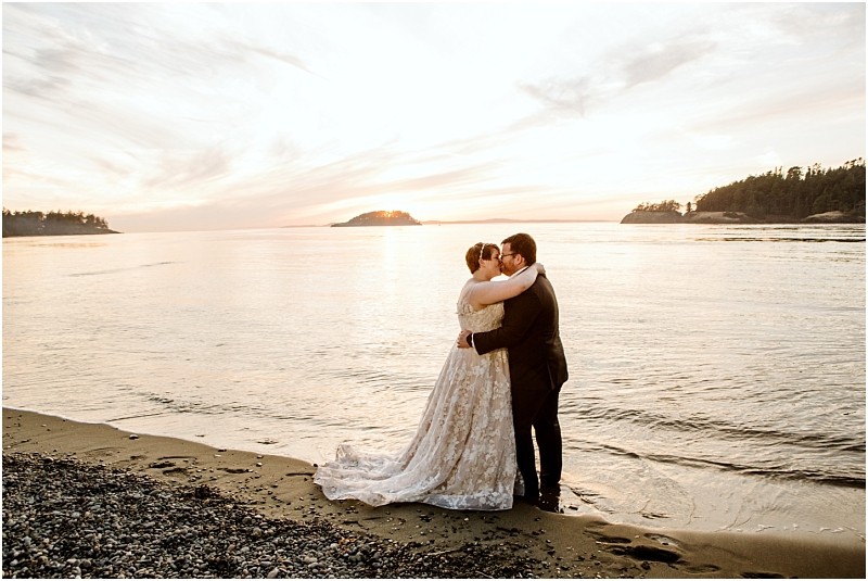 A bride and groom embrace and kiss on a snohomish beach at sunset with gentle waves and distant trees silhouetted against the sky The bride wears a long flowing gown and the groom is dressed in a dark suit Snohomish wedding photography
