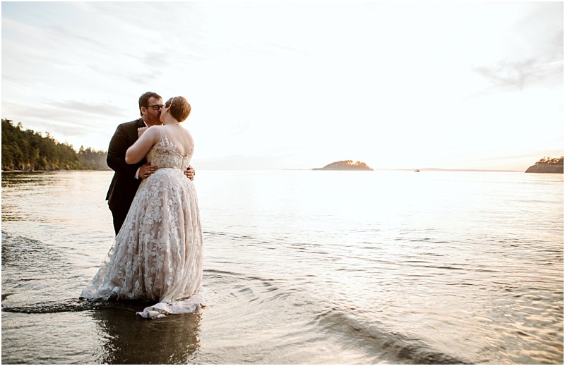 A couple in wedding attire stands in shallow water at the edge of a calm snohomish lake embracing and kissing at sunset with forested islands visible in the background Snohomish wedding photography