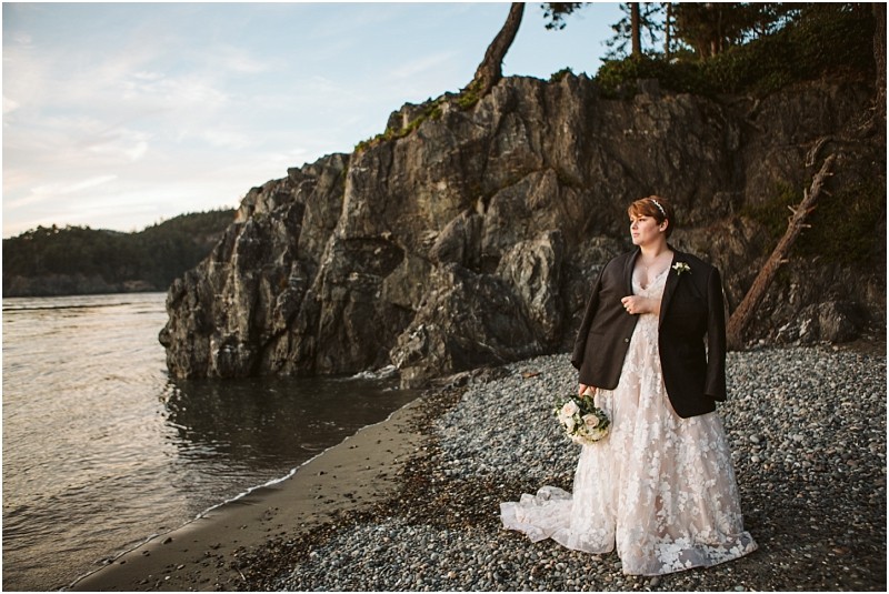A bride in a lace wedding dress and dark blazer stands on a rocky snohomish beach holding a bouquet with cliffs and calm water in the background at sunset Snohomish wedding photography