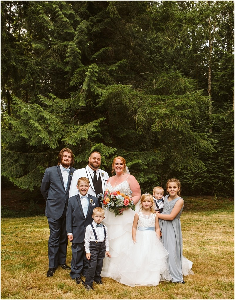 A wedding party poses outdoors on grass with tall green trees in the background capturing the charm of snohomish The bride in a white dress holds a bouquet beside the groom in a white jacket with four children and two adults smiling around them Snohomish wedding photography