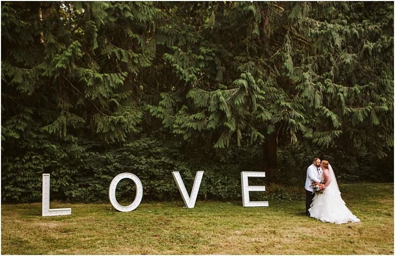 A bride and groom embrace and kiss outdoors in snohomish in front of large white letters spelling love with tall green trees in the background Snohomish wedding photography