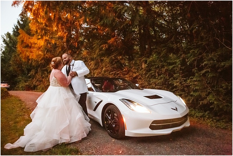 A bride and groom in formal attire stand close together beside a white sports car on a forested snohomish road at sunset with trees bathed in warm golden light Snohomish wedding photography