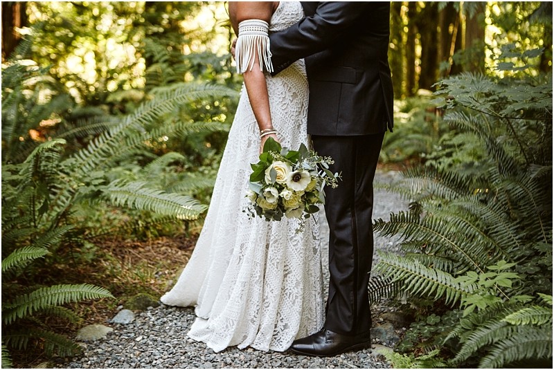 A bride in a lace gown holding a bouquet stands closely embraced by a groom in a black suit on a forest path surrounded by snohomish green ferns Their faces are not visible Snohomish wedding photography A bride in a lace gown holding a bouquet stands closely embraced by a groom in a black suit on a forest path surrounded by snohomish green ferns Their faces are not visible Snohomish wedding photography