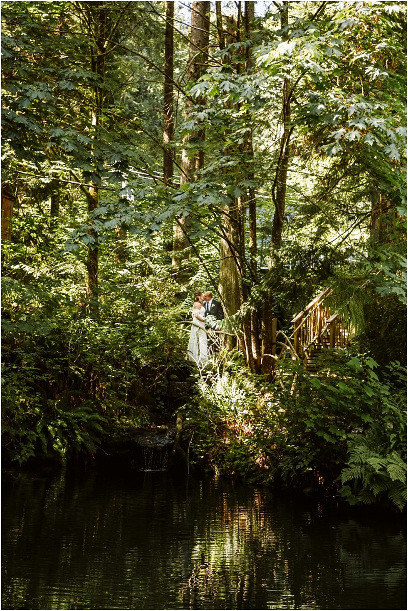 A bride and groom stand together on a small wooden bridge in snohomish surrounded by lush forest greenery with sunlight filtering through the trees and reflecting off a tranquil pond below Snohomish wedding photography