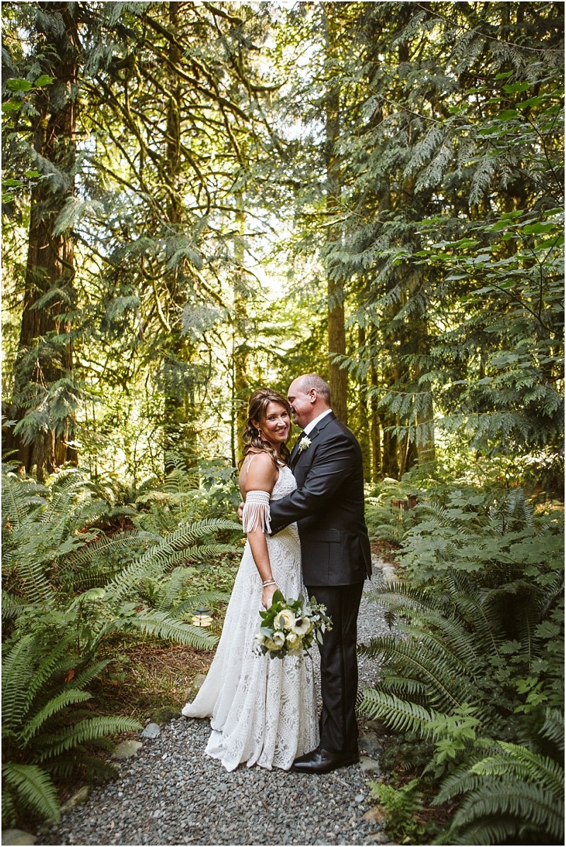 A bride in a white dress and a groom in a black suit embrace on a forest path in snohomish surrounded by lush green ferns and tall trees with sunlight filtering through the foliage Snohomish wedding photography