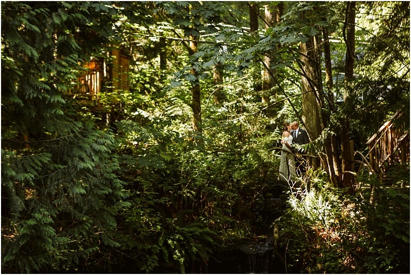 A couple stands close together in a sunlit snohomish forest surrounded by lush green trees and plants partially hidden among the foliage Dappled sunlight filters through the canopy creating a serene intimate atmosphere Snohomish wedding photography