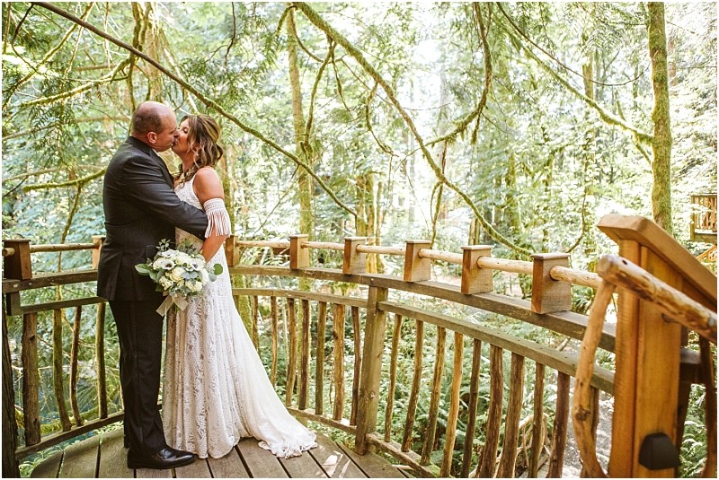 A bride and groom kiss on a wooden balcony in snohomish surrounded by lush green forest with sunlight filtering through the trees The bride holds a bouquet and wears a white dress while the groom is in a dark suit Snohomish wedding photography