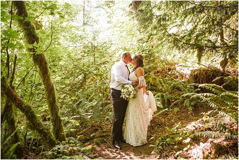 A bride and groom share a kiss in a lush sunlit snohomish forest The bride holds a bouquet and wears a white dress while the groom is dressed in a white shirt and dark pants Ferns and green foliage surround them Snohomish wedding photography