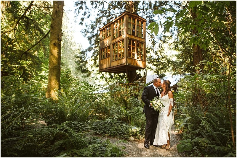 A bride and groom embrace on a forest path beneath a rustic wooden treehouse in snohomish surrounded by lush green foliage and tall trees Snohomish wedding photography