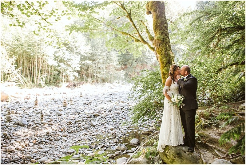 A bride and groom stand on a rock by a rocky snohomish riverbank in a sunlit forest embracing and smiling at each other The bride holds a bouquet and wears a white dress the groom wears a dark suit Snohomish wedding photography