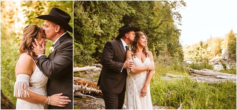 A couple dressed in wedding attire poses outdoors by a riverside in snohomish with fallen logs and lush greenery The man in a black suit and cowboy hat kisses the woman who wears a white lace dress on the forehead Snohomish wedding photography