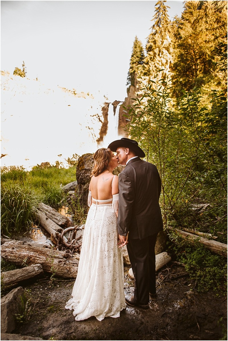 A bride in a white lace dress and a groom in a cowboy hat and suit stand hand in hand sharing a kiss outdoors by a snohomish waterfall surrounded by greenery and logs Snohomish wedding photography