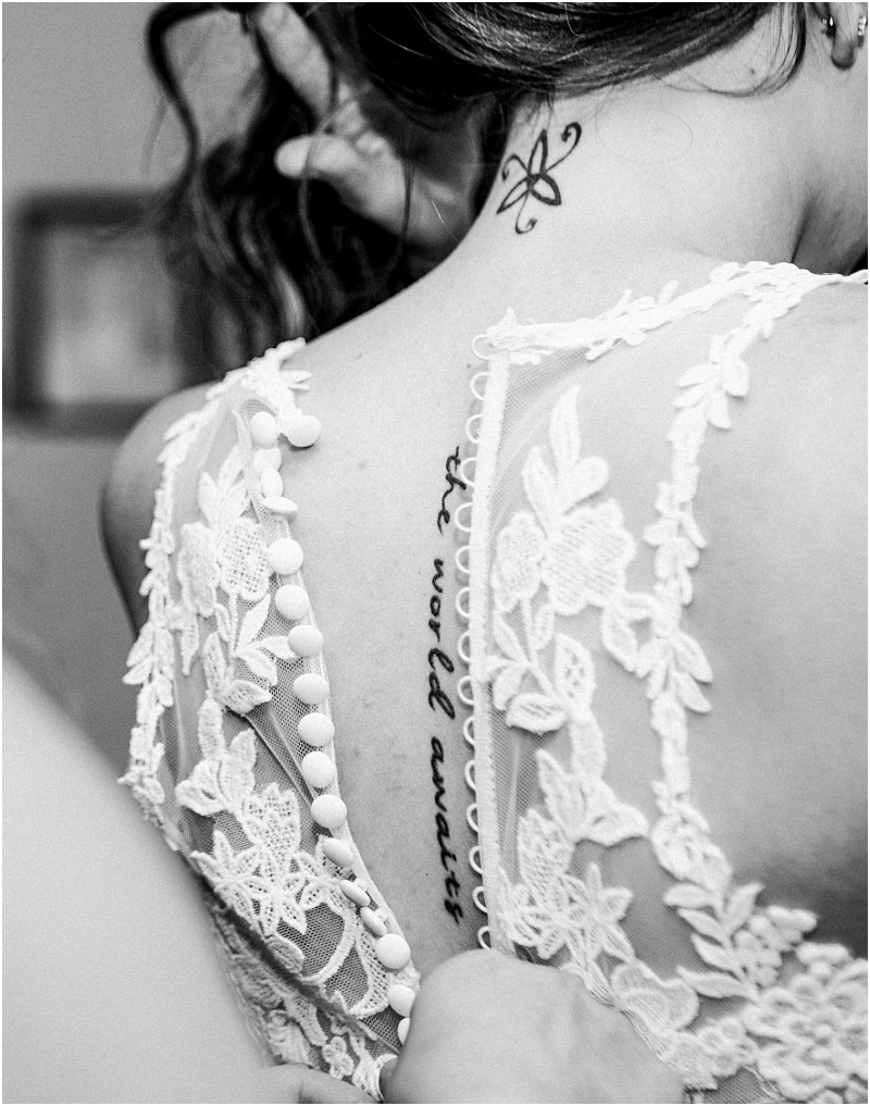 A close up black and white photo in snohomish captures a woman in a lace dress her back revealed with a text tattoo along her spine and a small atom symbol on her neck as another persons hand fastens the dress buttons Snohomish wedding photography