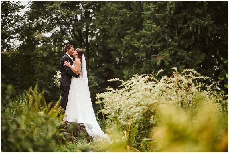A bride and groom stand on a stone ledge in snohomish embracing and kissing outdoors surrounded by lush greenery and tall trees The bride wears a long white dress and veil while the groom is in a dark suit Snohomish wedding photography