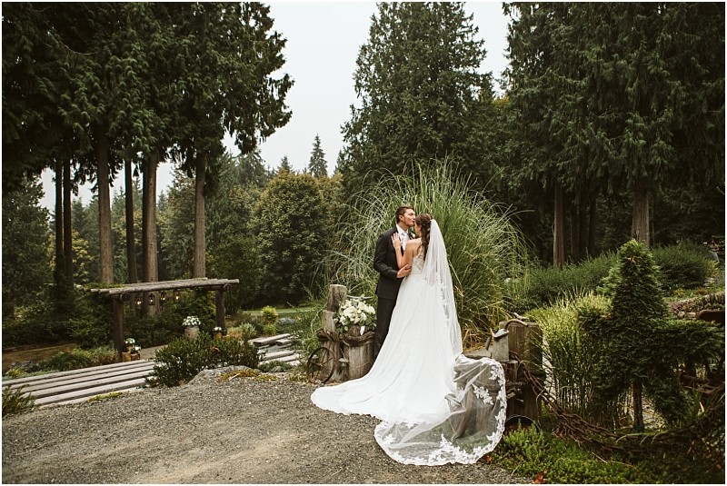 A bride and groom share a kiss outdoors in snohomish surrounded by lush greenery and tall trees The bride wears a long white gown and veil while the groom is in a dark suit amid rustic wooden benches and floral arrangements Snohomish wedding photography