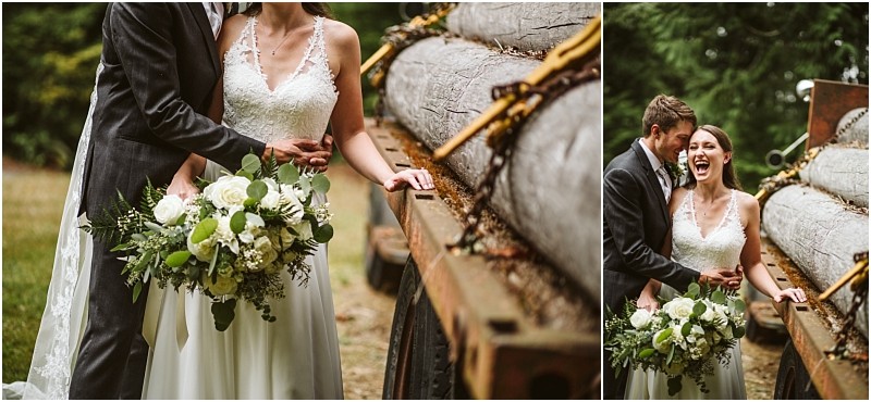 A bride in a white dress holds a bouquet of white flowers and greenery laughing with her groom in a dark suit beside a rustic log trailer outdoors in snohomish Snohomish wedding photography
