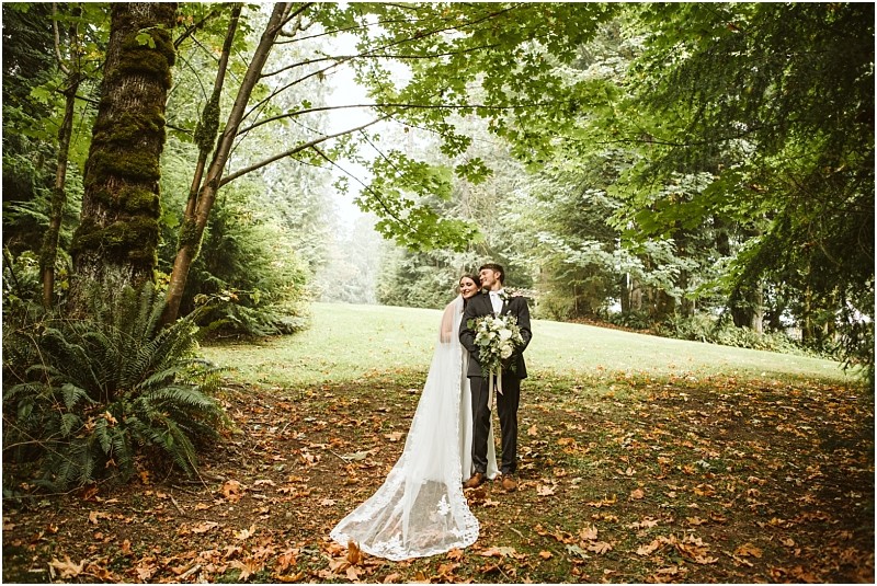 A bride and groom stand under lush green trees in a forested snohomish area with the bride in a long white dress and veil holding a bouquet Fallen leaves cover the ground creating a romantic natural setting Snohomish wedding photography